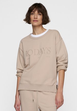Beige sweatshirt with ribbed cuffs and hem, featuring "10 DAYS" text on the front. Soft material, relaxed fit, side slits, layered white neckline.