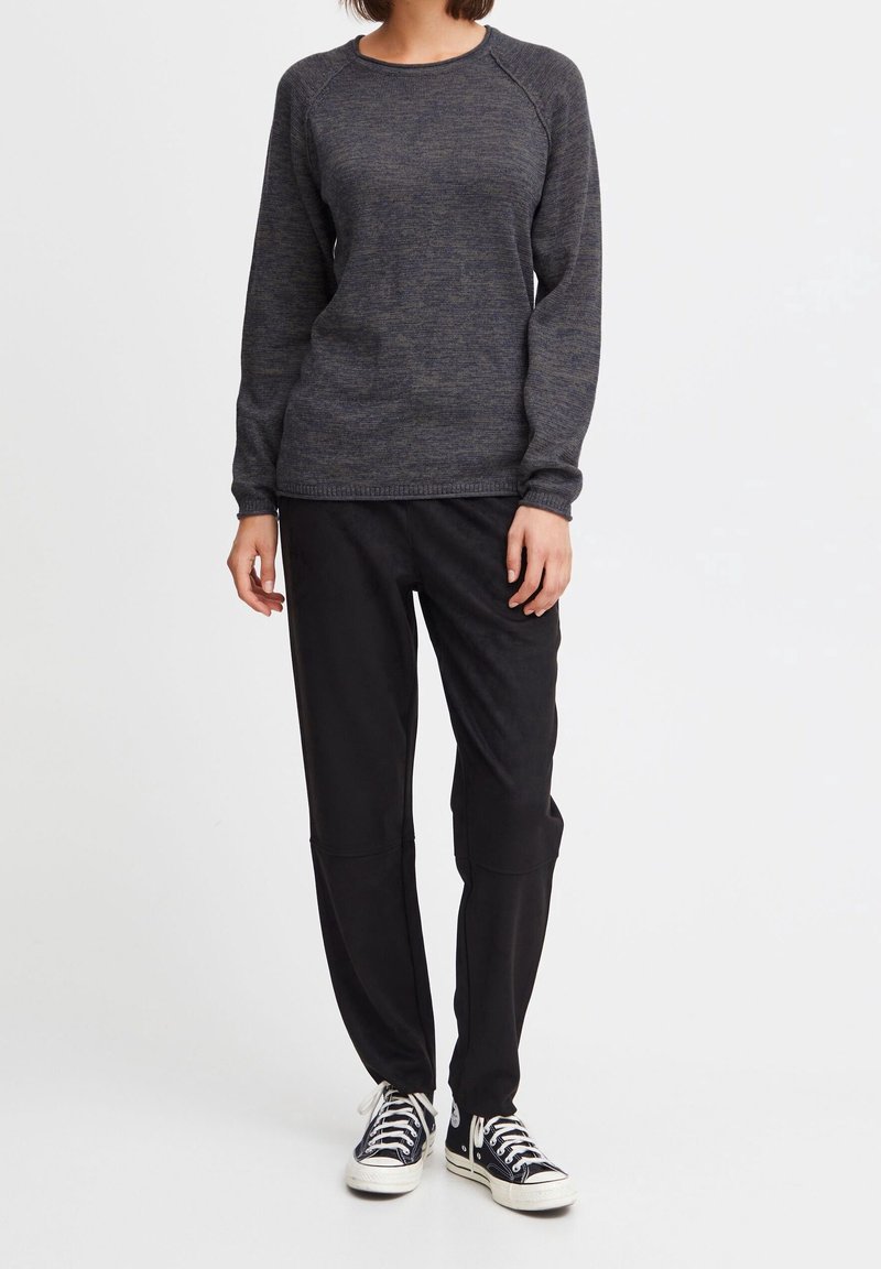 Personne portant un pull gris foncé à manches longues, un pantalon noir et des baskets basses noires avec des semelles et des lacets blancs, debout devant un fond blanc.