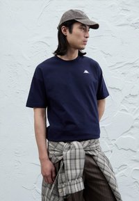 Marinblå t-shirt med kort ärm i bomull med en vit logotyp på bröstet. Modellen bär rutigt material knutet runt midjan.