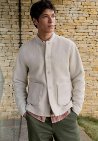 Light grey fleece jacket with a round neckline, front button closure, and two side pockets, layered over a plaid shirt and olive green pants.