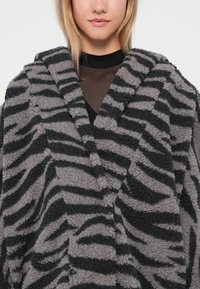 Étole en polaire à rayures grises et noires, présentant une texture douce, un large col et un design à recouvrement à l'avant, s'étendant jusqu'au niveau du milieu de la poitrine.