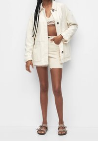 Veste en jean couleur crème avec deux poches avant, associée à un short crème assorti et des sandales marron. Haut en maille texturée sous la veste.