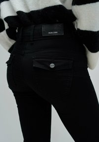 Jeans de mezclilla negra que presentan bolsillos traseros prominentes, una etiqueta de cuero con el logotipo "Salsa Jeans" y un sutil detalle de costura.