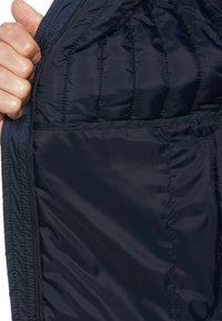 Marineblau gesteppte Jacke mit einem glatten Nylonfutter. Sie hat ein vertikales Steppmuster und eine Tasche auf der linken Seite.