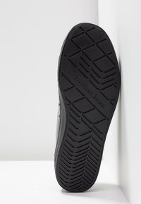 Semelle de basket noire avec motif géométrique de la bande de roulement et marquage en relief « Calvin Klein Jeans » au centre, photographiée sur fond blanc.