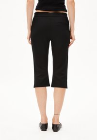 Zwarte cropped broek met een gladde textuur, een nauwsluitende taille en subtiele achterzakdetail. Lengte tot halverwege de kuit. Draagt met platte zwarte schoenen.