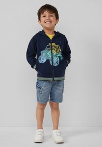 Marineblaue Zip-Up-Hoodie mit buntem Off-Road-Fahrzeug-Design, kombiniert mit hellen Denim-Shorts und weißen Sneakers. Jugendgröße, lässige Passform.