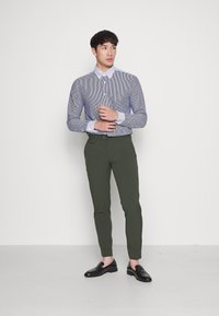Randig skjorta med lång ärm, kombinerad med olivgröna slim-fit byxor och svarta loafers, framför en neutral bakgrund.