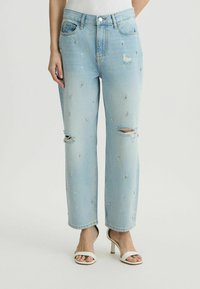 Jeans de color azul claro con un acabado desgastado, adornados, con rodilleras rasgadas, un corte recto y una cinta estándar en la cintura. Combinados con sandalias de tacón blancas.