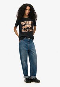 Camiseta de algodón negra con gráfico "SUPERDRY BLACK CATS" en oro rosa, combinada con jeans de mezclilla de tiro alto en azul claro y zapatos negros.