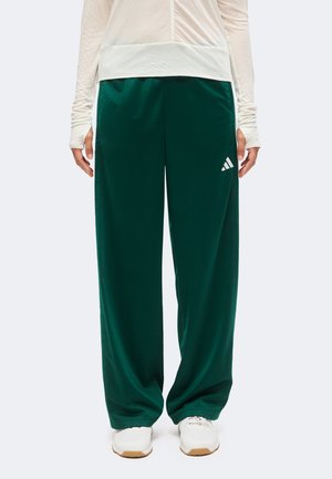 Person iført mørkegrønne Adidas træningsbukser med hvidt logo, hvide sneakers og en lys langærmet top, stående mod en ensfarvet baggrund.