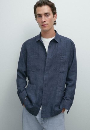 Dark blue button-up shirt made of textured fabric with two chest pockets and a relaxed fit, paired with a light undershirt and denim jeans.