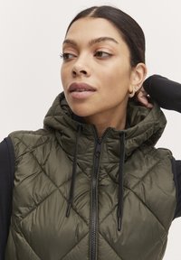 Gilet matelassé vert olive avec capuche, disposant d'une fermeture éclair à l'avant, de cordons noirs et de motifs en losange sur le tissu.