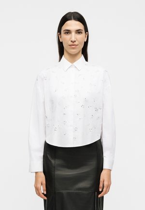 Femme aux cheveux lisses et foncés portant une chemise blanche à manches longues avec broderie florale et une jupe en cuir noire, debout face à nous.