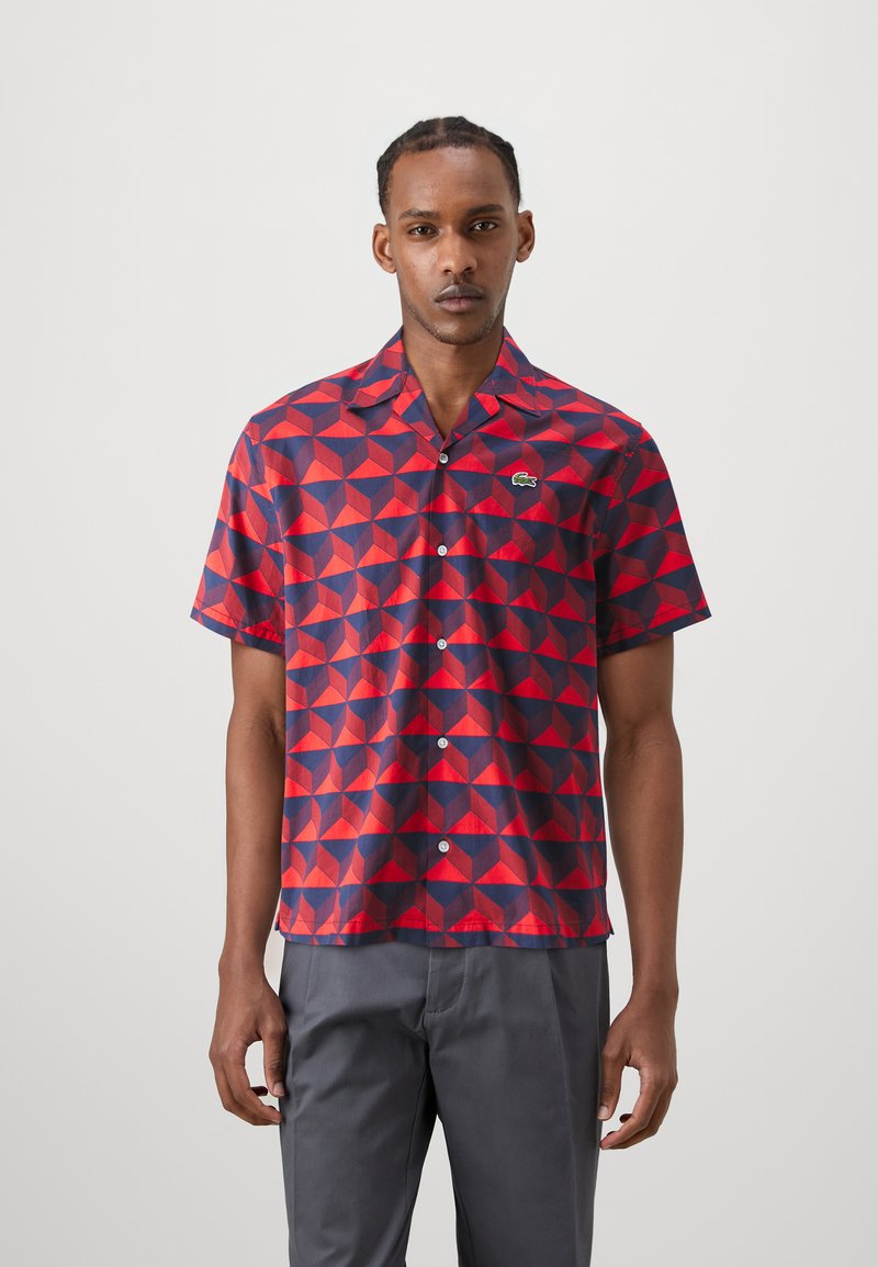 Chemise à manches courtes avec un motif géométrique rouge et bleu, fabriquée en tissu doux ; elle se ferme par des boutons à l'avant et présente un petit logo brodé sur la poitrine.