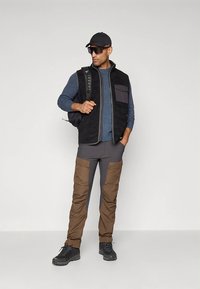 Svart fleecefodrad väst med en lila bröstficka, kombinerad med grå smala byxor och bruna cargo-accenter. Svarta vandringskängor och keps.