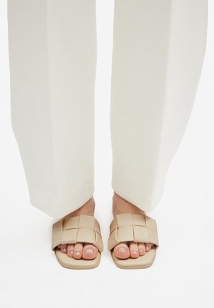 Beżowe klapki na płaskim obcasie z plecionym paskiem, kwadratowymi noskami i gładkim wykończeniem, noszone z luźnymi białymi spodniami.