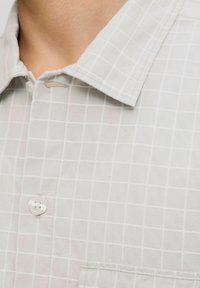 Primo piano di una camicia bianca con colletto, caratterizzata da un motivo a griglia quadrata, che mostra il colletto, un bottone bianco e parte di una tasca sul petto.