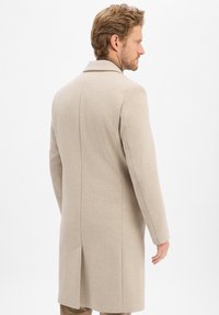 Man met lichtbruin haar in een beige lange jas, staande en kijkend naar de zijkant tegen een effen witte achtergrond.