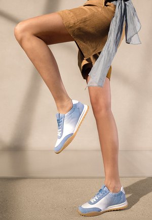 Legs wearing brown shorts and blue sneakers, one foot lifted mid-step against a plain beige background.