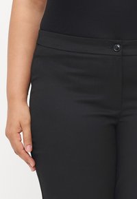 Black tailored pants with a smooth texture, featuring a front button closure and a slim fit design, visible hand placement on the waistband.