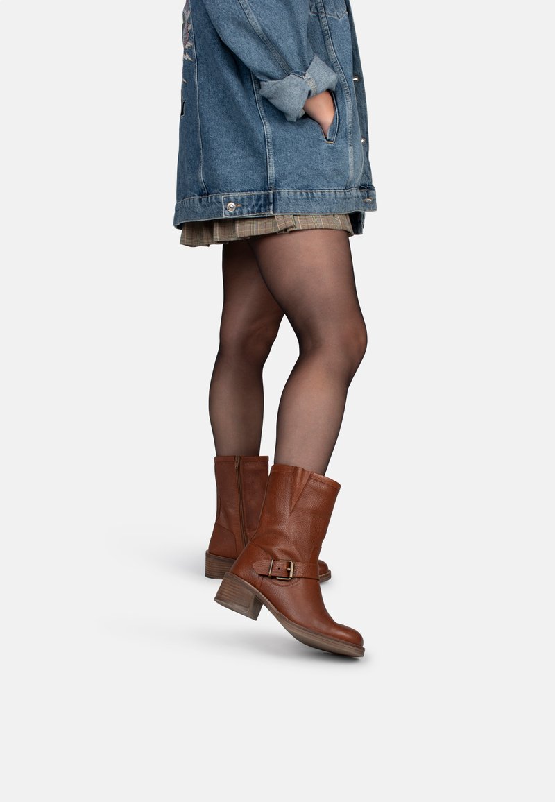 Bottines en cuir marron avec un talon épais, dotées d'une fermeture éclair latérale et d'une boucle décorative. Portées avec une veste en denim et une jupe à carreaux.