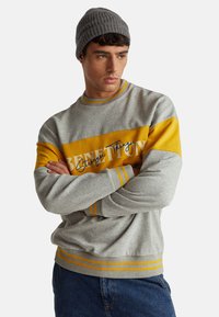 Uomo con felpa Benetton grigia e gialla e berretto di maglia grigio, braccia incrociate, guarda di lato contro uno sfondo semplice.