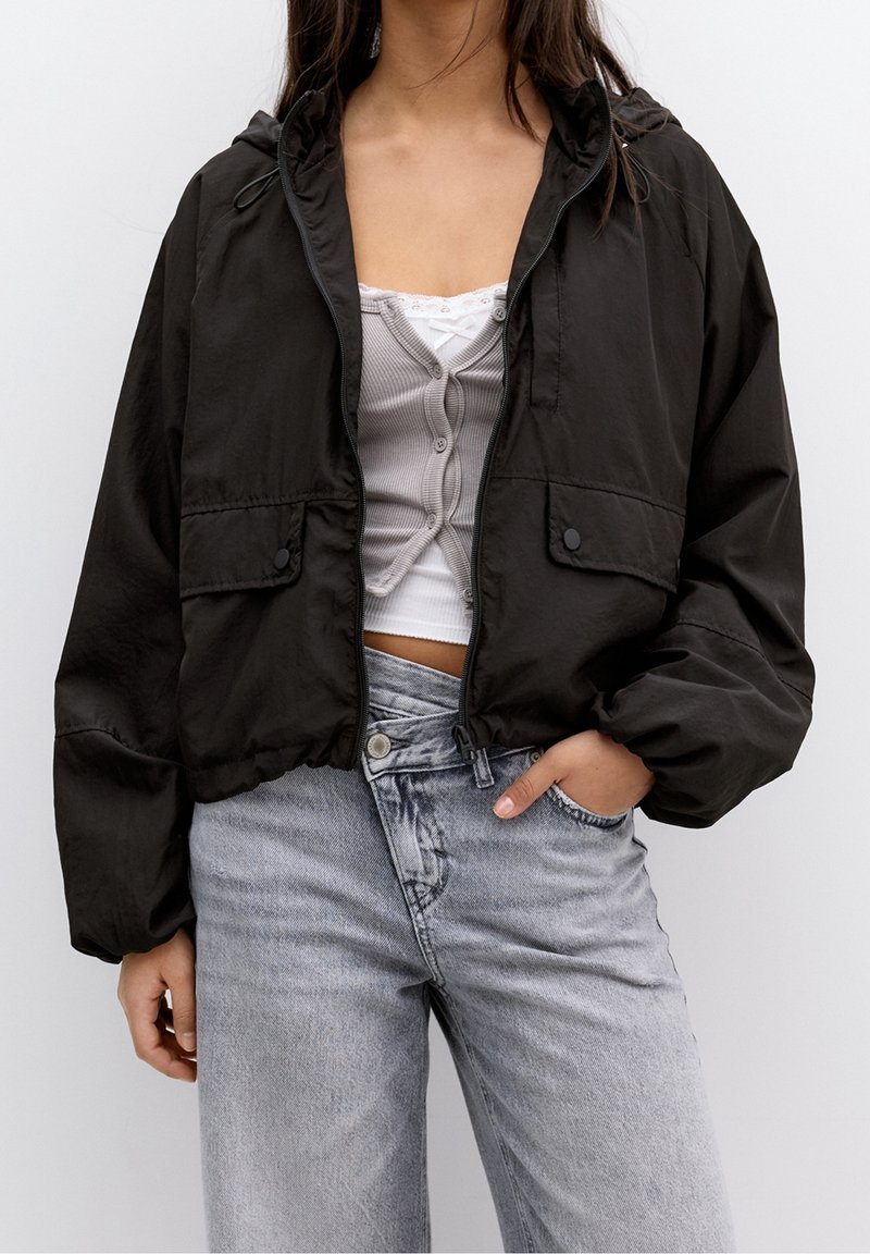 Femme portant une veste courte noire par-dessus un cardigan gris boutonné et un haut blanc avec un jean taille haute gris clair.