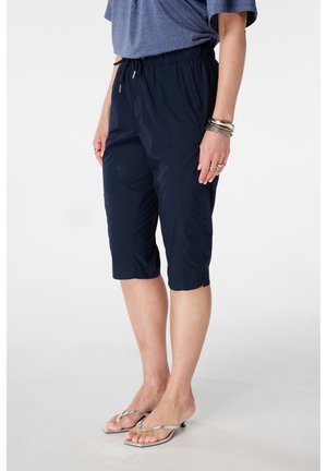 Personne portant un short bleu marine jusqu'aux genoux et des tongs argentées à talons, debout, les mains détendues le long du corps sur un fond blanc.