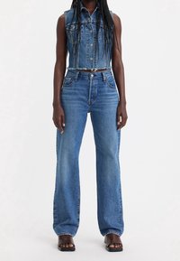 Mujer con chaqueta de mezclilla sin mangas y corta, pantalones vaqueros de tiro alto y pata ancha en azul, y sandalias de tacón abiertas negras sobre fondo blanco.