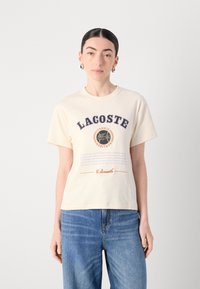 Majica kratkih rukava krem boje s podebljanim natpisom "LACOSTE", grafikom tenisa i tiskanim detaljima. Uparena s svijetloplavim trapericama.