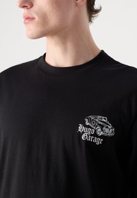 Black cotton t-shirt featuring a white graphic of a vintage car and the text “Hugo Garage” on the left chest area. Crew neck design.
