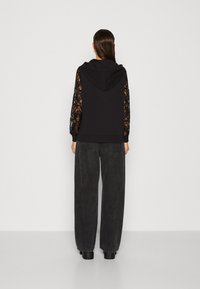 Sudadera negra con detalles de encaje en las mangas, con capucha y un corte relajado. Combinada con unos vaqueros oscuros de pierna ancha, creando un atuendo casual.