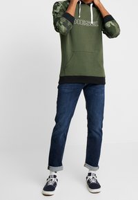 Zielona bluza z kapturem z kamuflażowymi rękawami, białym logo, przednią kieszenią, zestawiona z ciemnoniebieskimi dżinsami z podwiniętymi mankietami oraz granatowymi sneakersami.