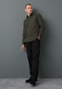 Camicia verde a quadri con bottoni e maniche lunghe, abbinata a pantaloni di jeans neri e scarpe nere, in piedi contro uno sfondo grigio.