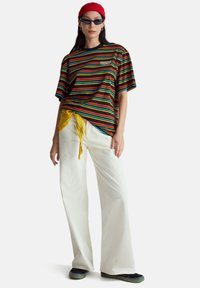 Pruhované tričko v červené, žluté, zelené a modré barvě s černým výstřihem; doplněné bílými džíny s širokými nohavicemi a žlutým šátkem uvázaným v pase.