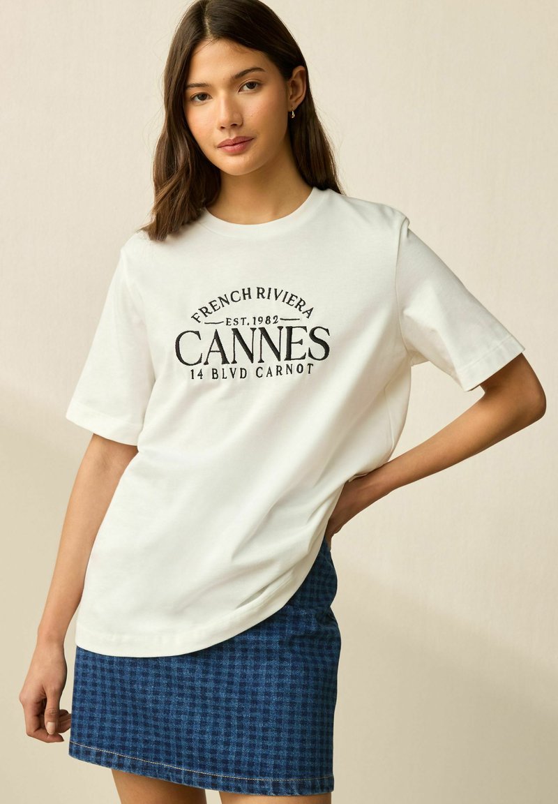 Weißes Baumwoll-T-Shirt mit kurzen Ärmeln, bedruckt mit "FRENCH RIVIERA CANNES" in schwarzer Schrift. Kombiniert mit einem karierten Jeansrock.
