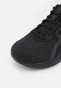 Chaussure de sport noire avec une tige en mesh texturé, un embout renforcé et une semelle à motifs, aux lignes épurées et au design minimaliste.