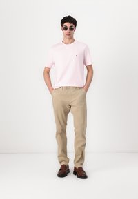 T-shirt en coton rose clair avec logo, pantalon beige et chaussures marron. Les accessoires incluent des lunettes de soleil noires. Le modèle se tient les mains dans les poches.