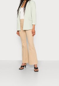 Blazer verde menta con una vestibilità rilassata, abbinato a pantaloni beige a zampa d'elefante e sandali neri con tacco, caratterizzati da una texture liscia e un design sottile.