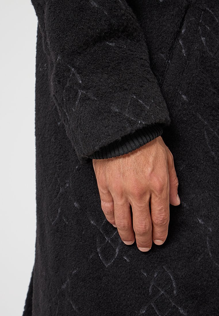 Manteau en laine noire avec une finition texturée. Présente des poignets côtelés et des détails en motif. La vue de côté montre une main reposant sur le tissu.