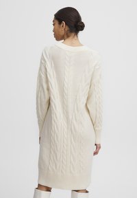 Cremefarbenes Strickkleid mit langen Ärmeln, geripptem Saum und kunstvollen Strukturmustern auf dem Rücken und den Ärmeln.