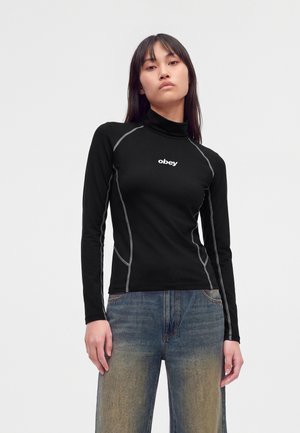 Jeune femme portant un col roulé noir à manches longues avec le logo "obey" et des coutures grises, associée à un jean bleu délavé, debout devant un fond blanc.
