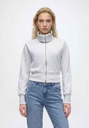Une femme blonde porte une veste blanche zippée à col montant et un jean bleu, sur un fond clair et uni.