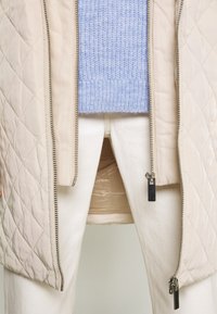 Gros plan sur une personne portant une veste matelassée beige avec des fermetures éclair doubles, un pantalon crème et un pull en tricot bleu clair.