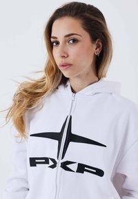 Sudadera blanca con cremallera y un logo negro que presenta una estrella estilizada y el texto "PXP". Tejido suave con un corte holgado y capucha forrada.