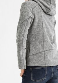 Tylny widok osoby w dopasowanej szarej kurtce z kapturem i ciemnoniebieskich dżinsach z widocznymi detalami przeszyć.