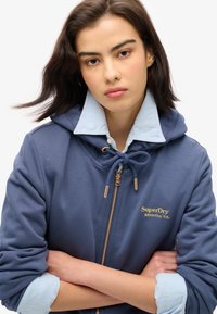Marineblå zip-up hoodie med en lyseblå krave. Har guld "SuperDry" logo, snøring i hætten og frontlynlås. Blød stofstruktur.