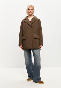 Chaqueta acolchada marrón con collar de solapa, dos bolsillos frontales y un corte relajado, combinada con vaqueros de pierna ancha de color azul claro y zapatos marrones.
