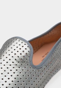 Ασημί επίπεδο παπούτσι με τρύπες, με απαλό περίγραμμα περιθωρίου και καφέ επένδυση με μαξιλάρι που φαίνεται στο εσωτερικό.